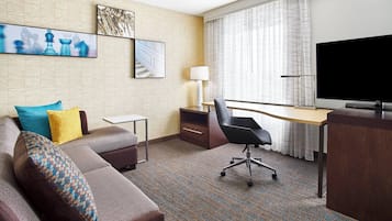 Habitación de hotel con sofá, escritorio, silla, televisión y obras de arte en las paredes.