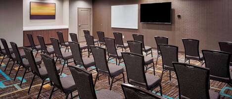 A conference room with rows of chairs, a whiteboard, and a flat-screen TV on the wall.
