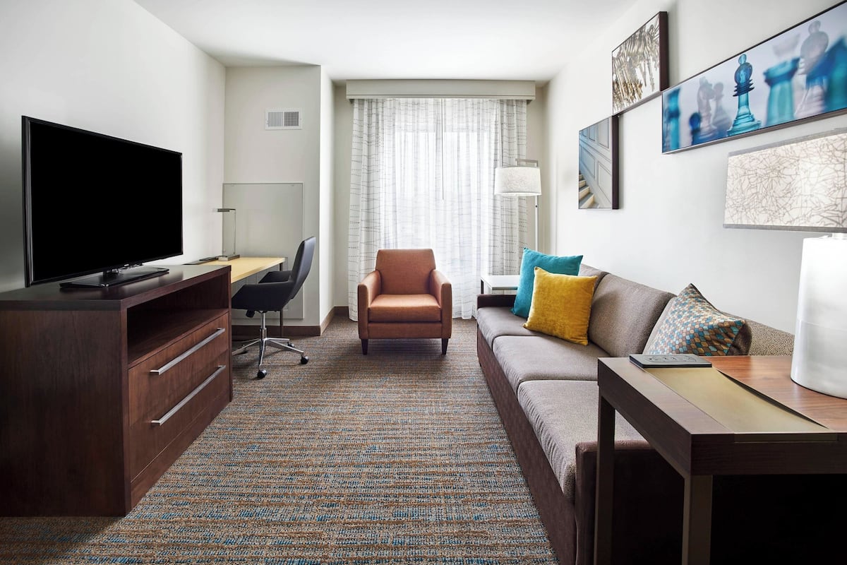 Une chambre d’hôtel équipée d’un téléviseur à écran plat, d’un bureau en bois, d’un fauteuil marron, d’un canapé agrémenté de coussins colorés et d’œuvres d’art accrochées aux murs.
