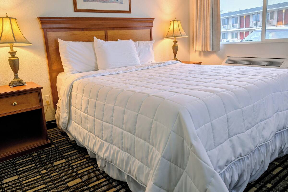 A neatly made bed with white bedding, two bedside lamps, a wooden headboard, and a framed picture on the wall.