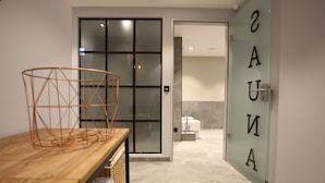 A modern bathroom with a glass door labeled "SAUNA," a wooden vanity with a basket, and a tiled floor.