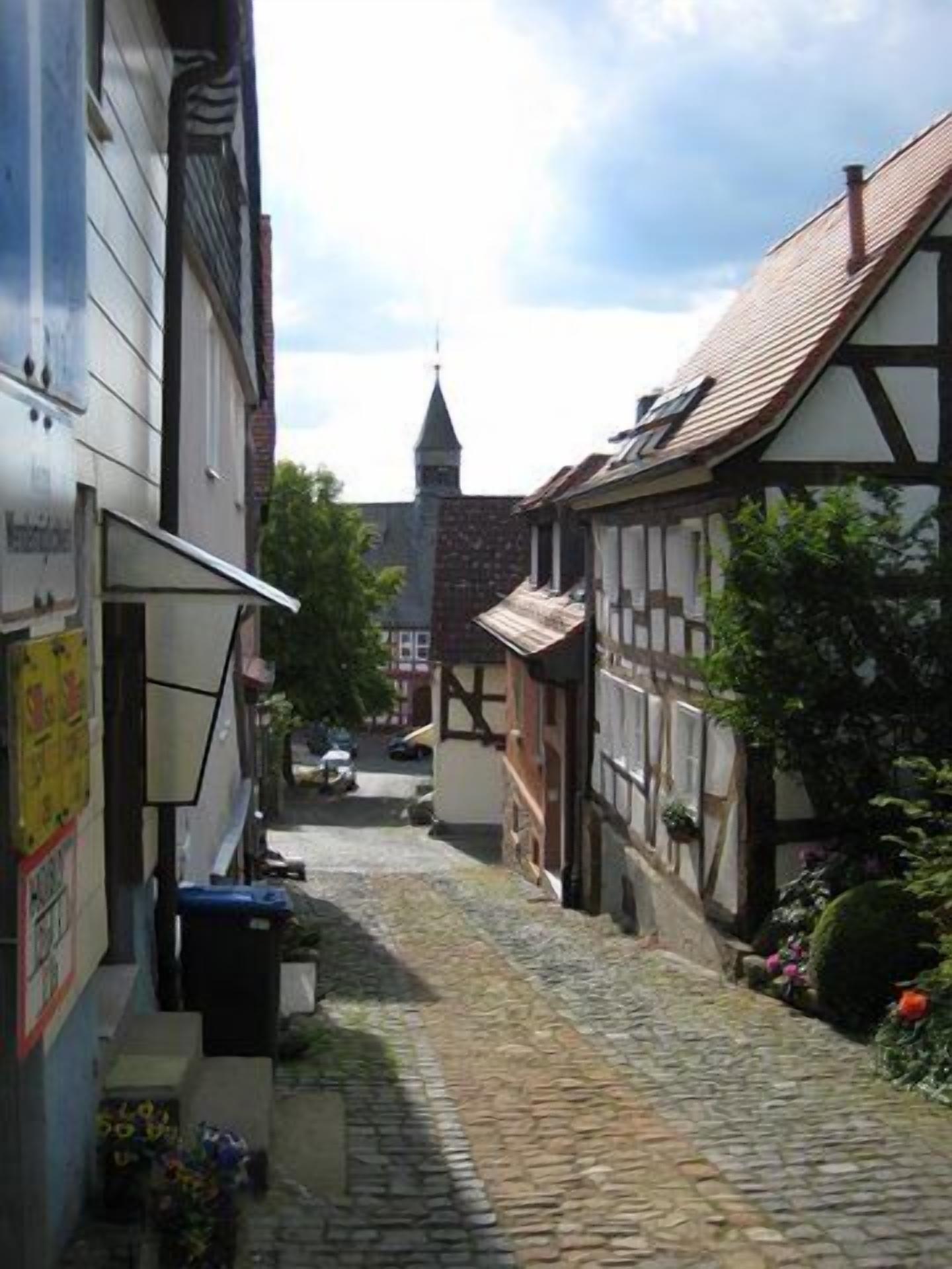 A narrow cobblestone street lined with traditional half-timbered buildings, a church steeple in the background, and a clear sky.