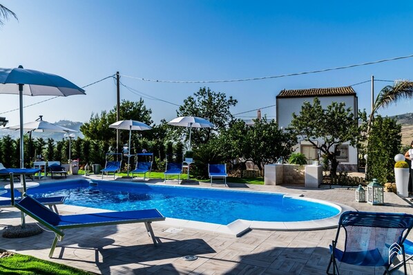 A swimming pool with lounge chairs and umbrellas, surrounded by greenery and a building with a tiled roof.