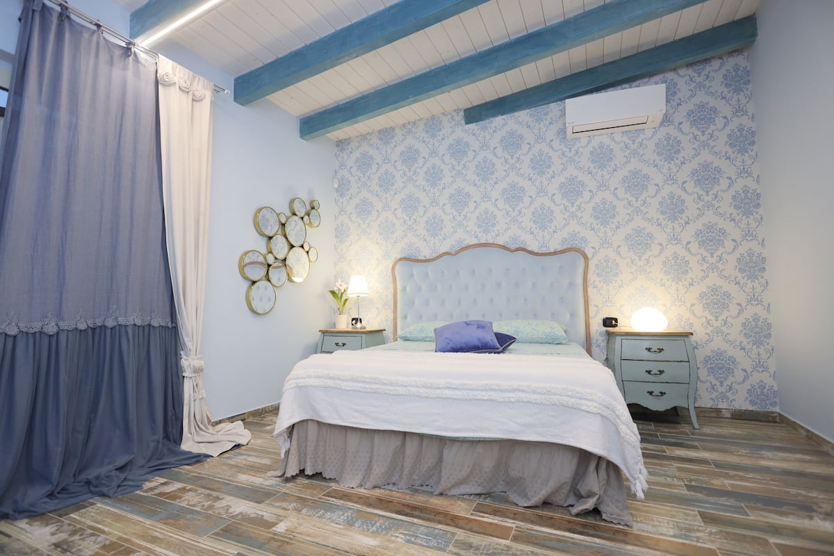 A bedroom with a bed, two bedside tables, a nightstand, a wall-mounted air conditioner, and patterned wallpaper.