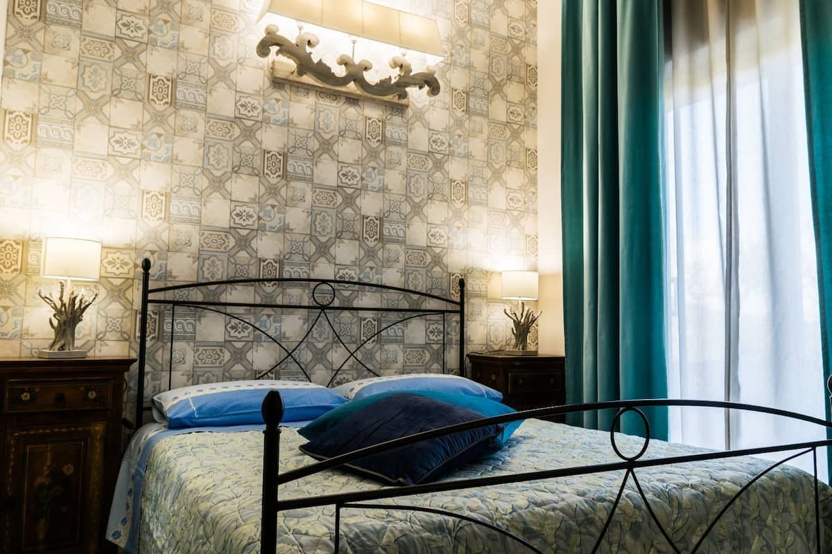 A bedroom with a metal bed frame, patterned wallpaper, two bedside lamps, and a window with curtains.