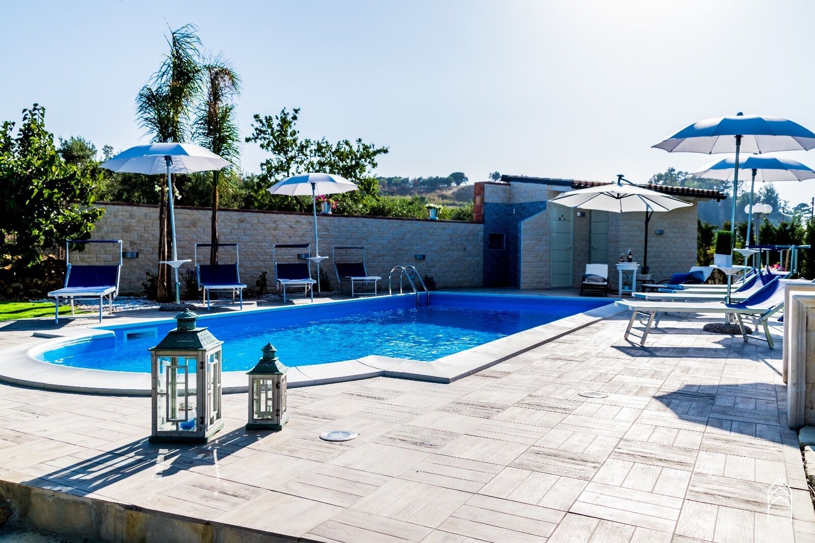 Une piscine entourée d’un mur de pierre et de verdure, avec des chaises longues et des parasols.