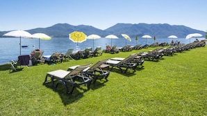 A row of sun loungers with umbrellas on a grassy area by a lake, with mountains in the background.
