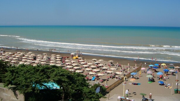 Uma praia com vários guarda-sóis e espreguiçadeiras, céu claro e mar calmo.