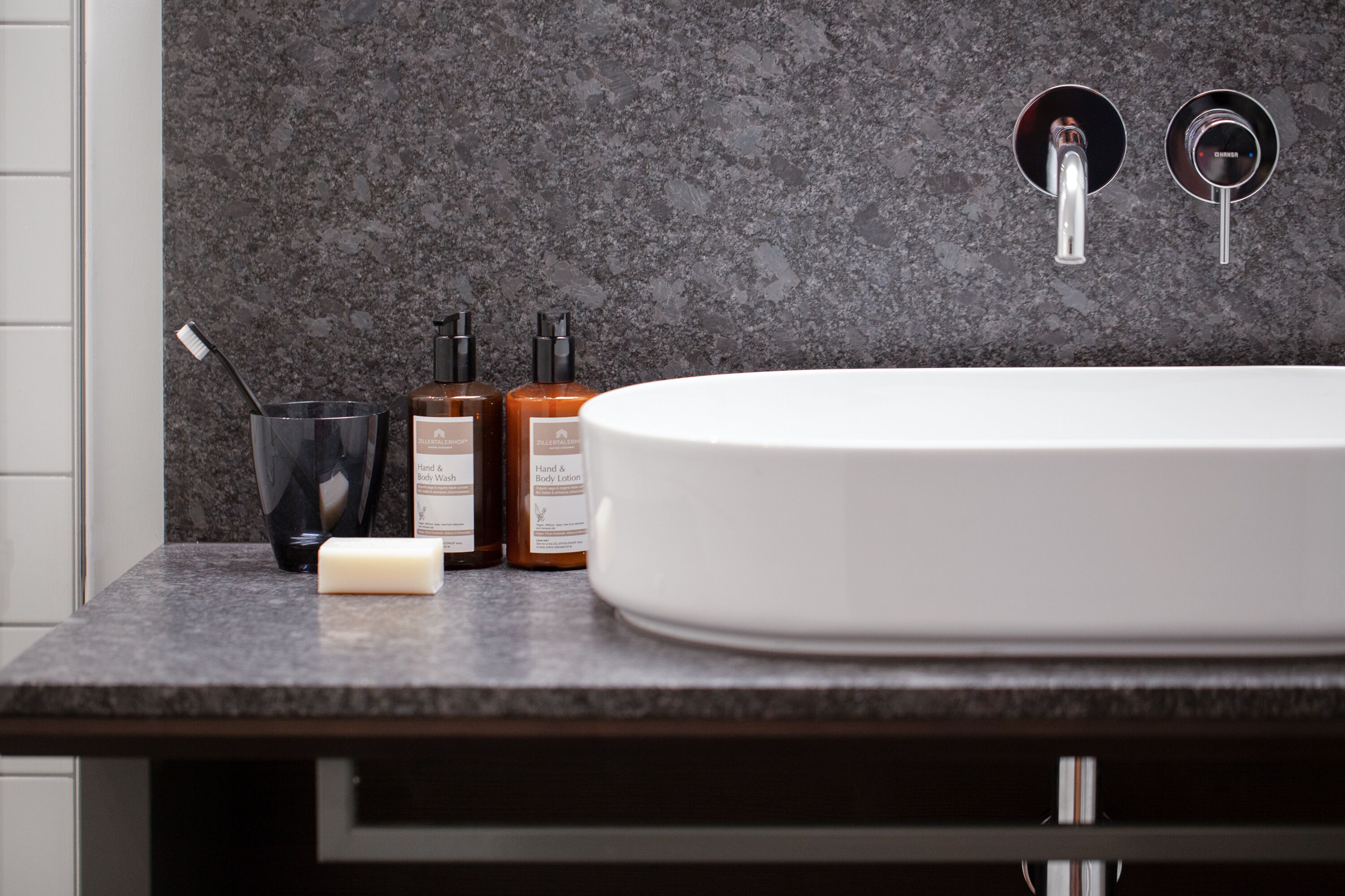 Un baño con un lavamanos blanco, encimera oscura y dos grifos. Hay tres botellas de jabón de manos y cuerpo sobre la encimera.