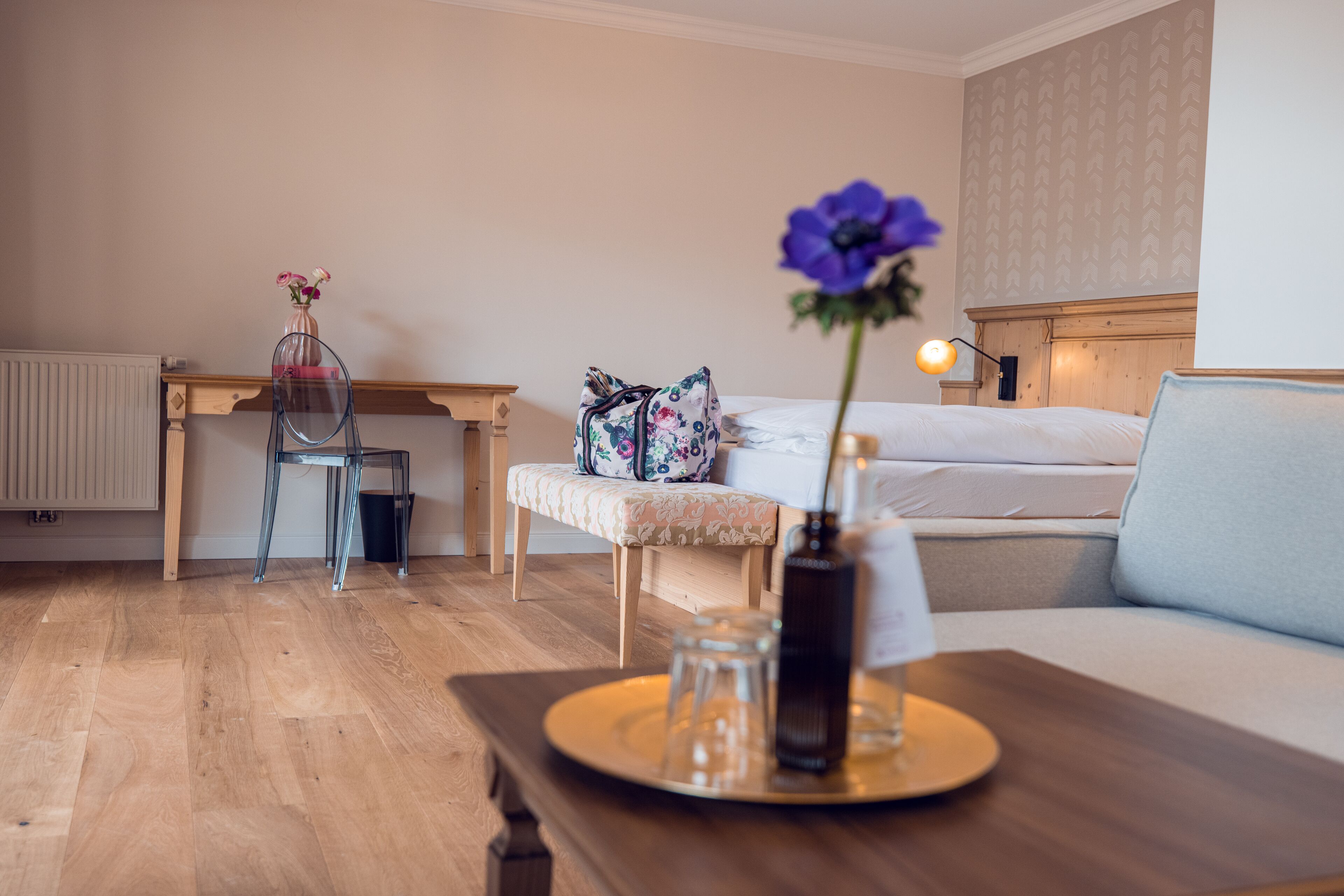 Una habitación de hotel acogedora con suelo de madera, una cama con ropa blanca, un sofá, una mesita con un florero y una vela, y un banco con cojín estampado.