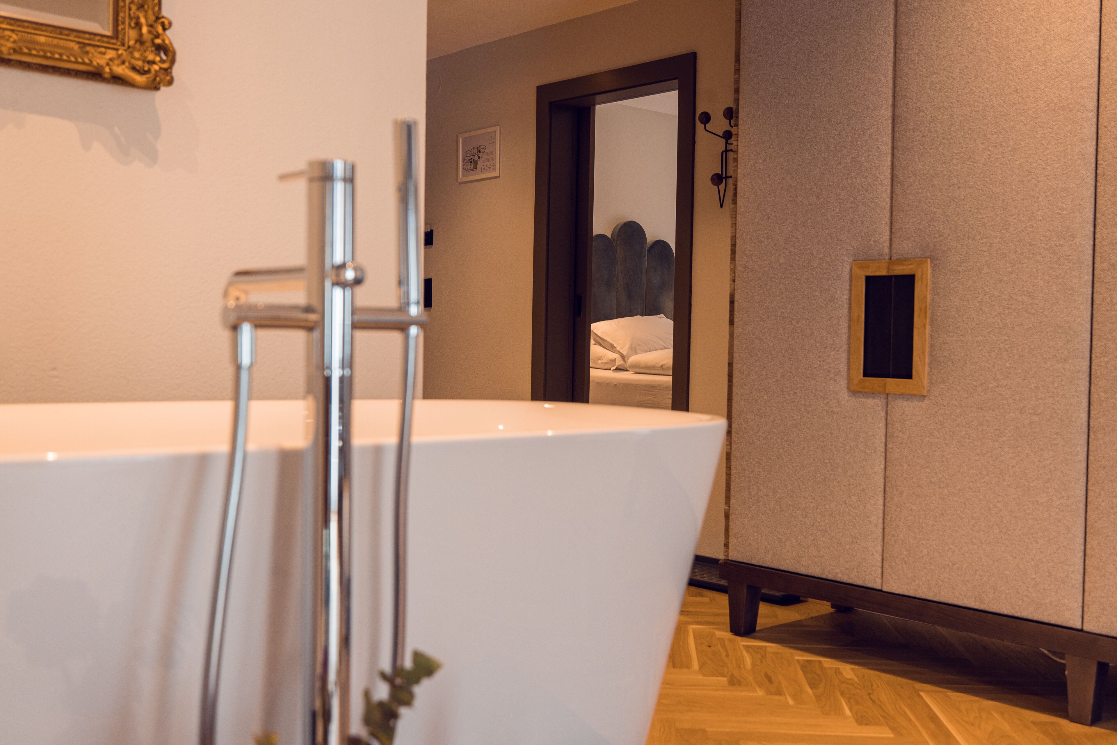 A modern bathroom with a freestanding bathtub, a wooden floor, and a mirror reflecting a bed.