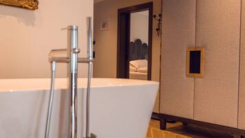 A modern bathroom with a freestanding bathtub, a wooden floor, and a mirror reflecting a bed.