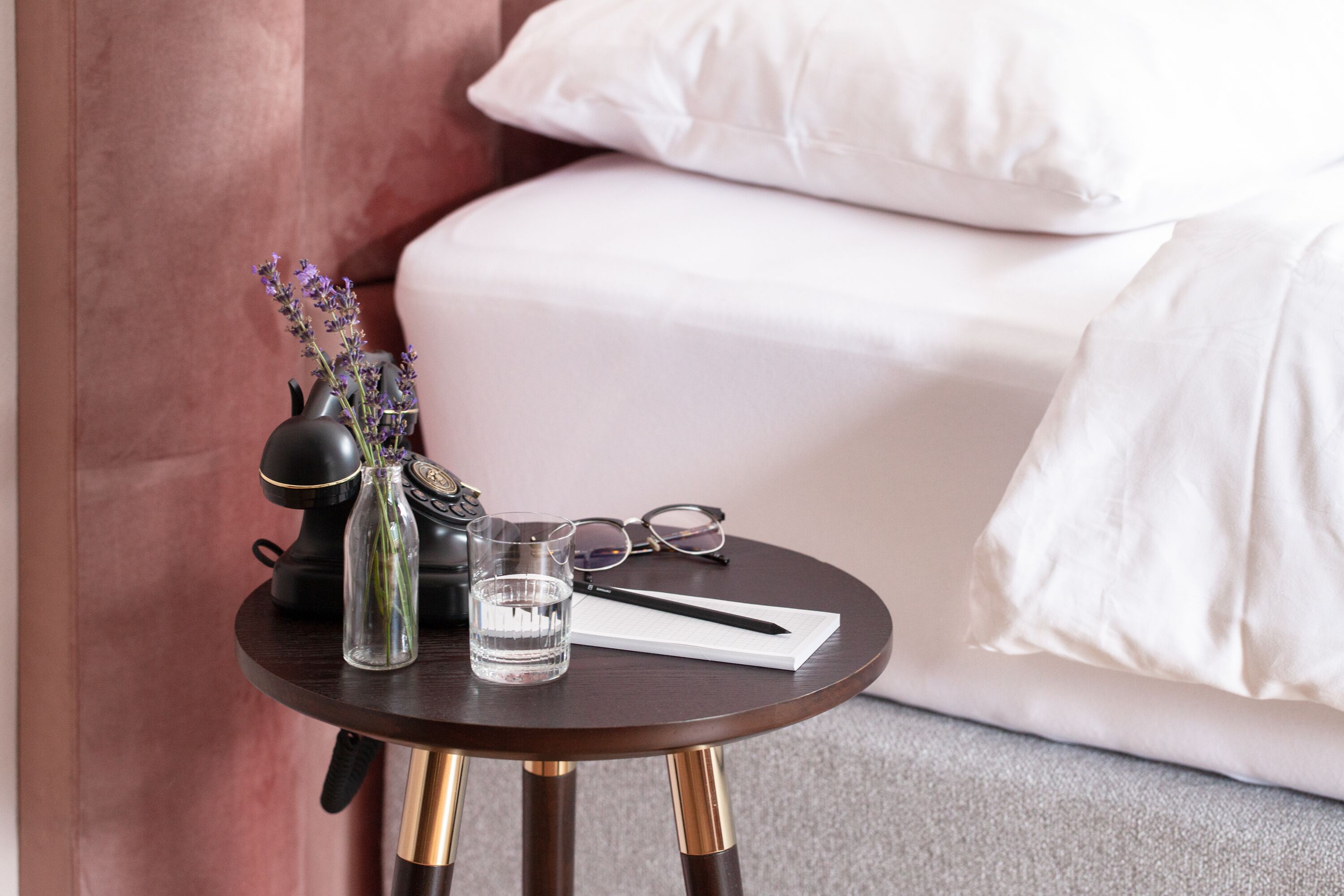 A small round table with a dark wood finish and gold legs, placed on a carpeted floor. On the table, there is a clear glass of water, a black vase with purple flowers, a pair of glasses, and a white notepad with a pen.