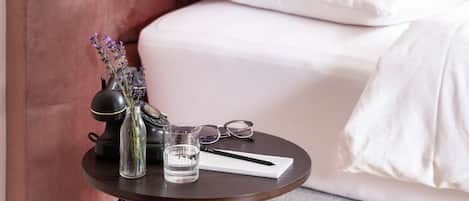Una pequeña mesa redonda con acabado en madera oscura y patas doradas, ubicada sobre un piso alfombrado. Sobre la mesa, se encuentra un vaso de vidrio transparente con agua, un florero negro con flores moradas, un par de gafas y una libreta blanca con un bolígrafo.