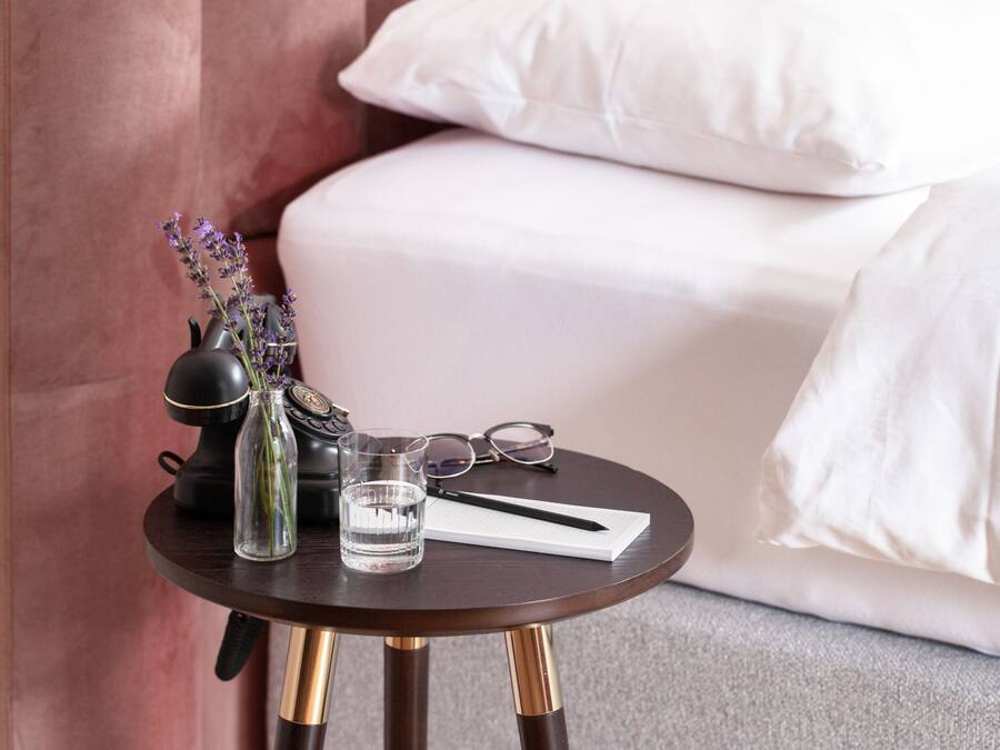 A small round table with a dark wood finish and gold legs, placed on a carpeted floor. On the table, there is a clear glass of water, a black vase with purple flowers, a pair of glasses, and a white notepad with a pen.