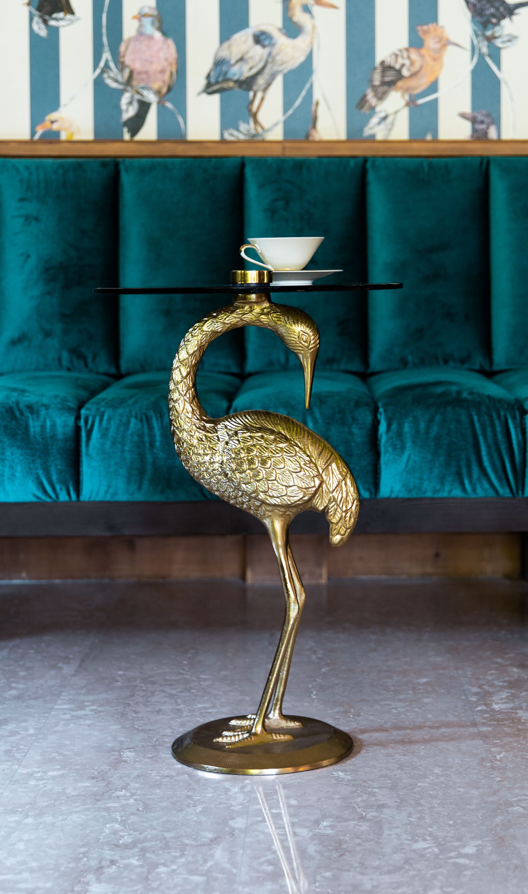 Una escultura dorada con forma de pájaro sobre un pedestal, una taza y platillo blancos encima, y un sofá tapizado color aguamarina al fondo.