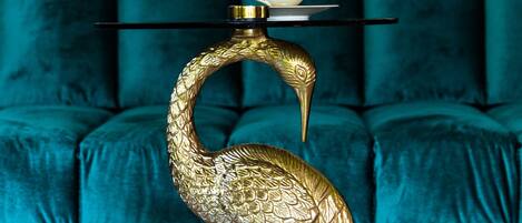 A golden bird-shaped sculpture on a pedestal, a white cup and saucer on top, and a teal upholstered sofa in the background.