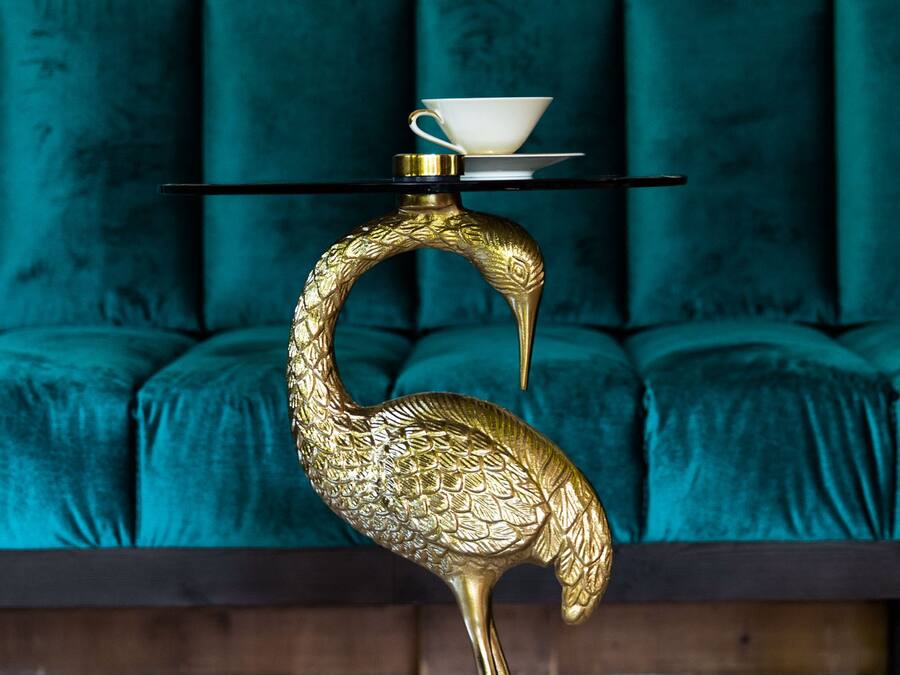 Una escultura dorada con forma de pájaro sobre un pedestal, una taza y platillo blancos encima, y un sofá tapizado color aguamarina al fondo.