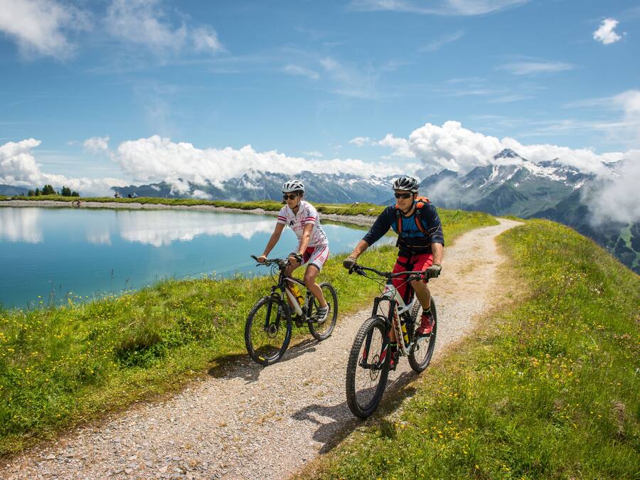 Dos ciclistas en un camino de grava junto a un lago, con montañas al fondo.