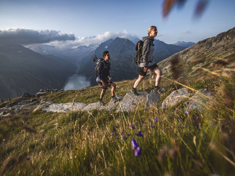 Dos excursionistas en un sendero de montaña, con un lago y montañas al fondo.