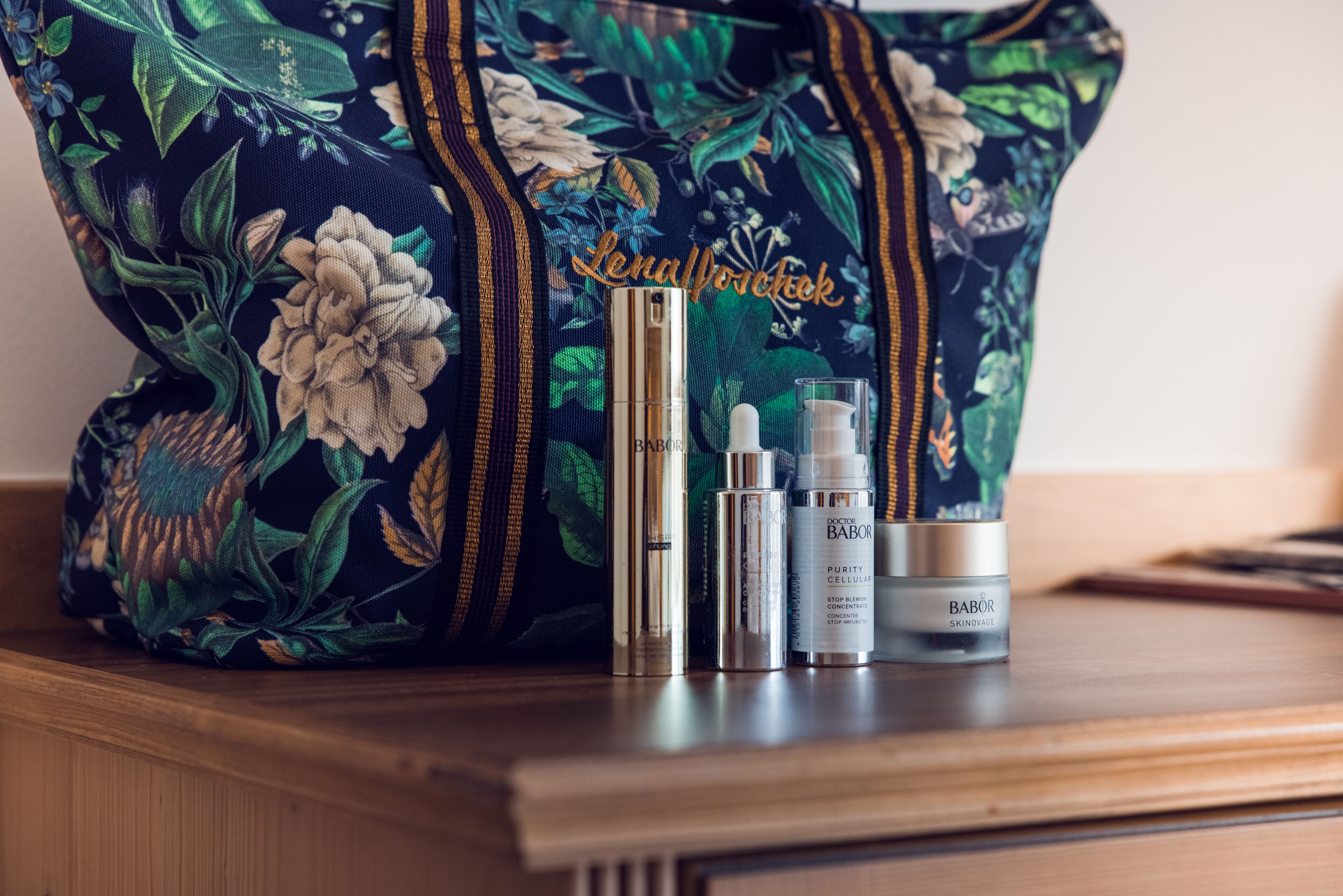 A floral-patterned bag, a gold-colored bottle, and two skincare containers on a wooden surface.