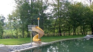 A playground with a spiral slide over a pool, surrounded by trees and a fenced area.