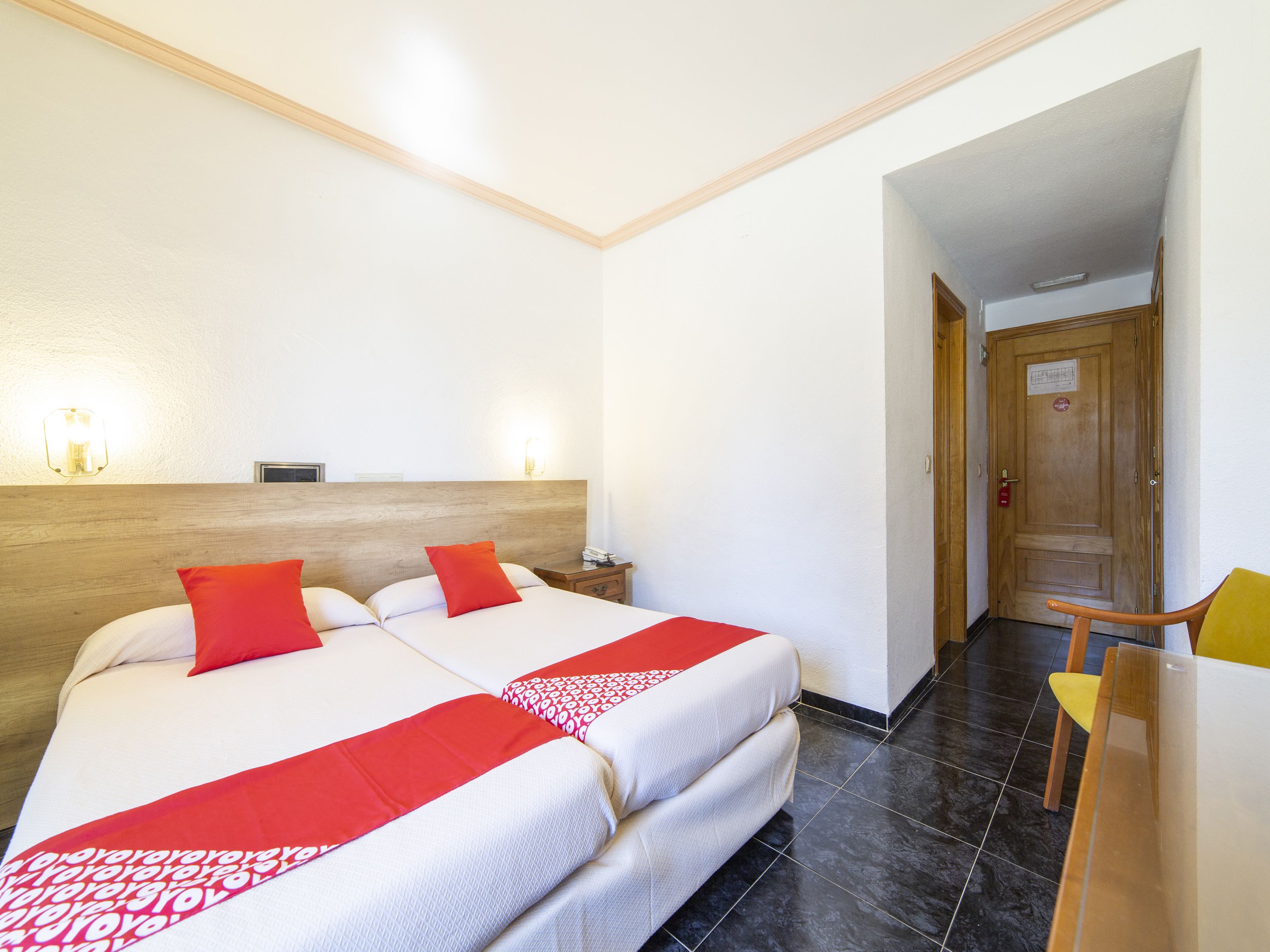 Habitación de hotel con cama doble, dos almohadas rojas, cabecera de madera, mesita de noche con lámpara y una silla con cojín amarillo.