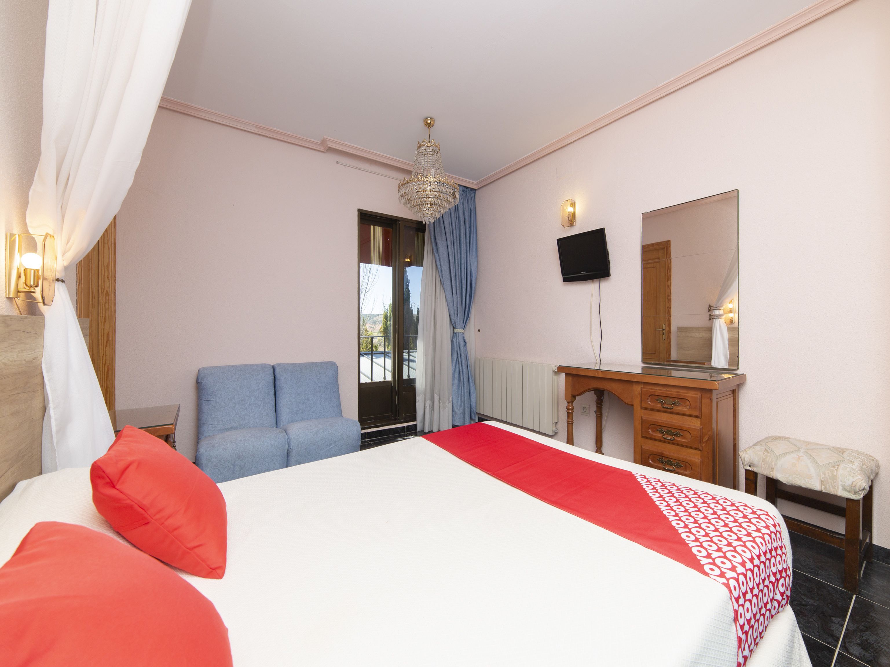 A hotel room with a bed, a desk, a chair, a TV, and a balcony with curtains.