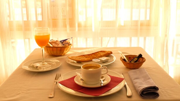 Una mesa de desayuno con un vaso de zumo de naranja, una taza de café, un plato de cruasanes, una cesta de pan y un bol de mantequilla.