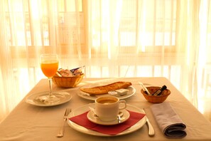 Une table de petit-déjeuner avec un verre de jus d’orange, une tasse de café, une assiette de croissants, un panier de pain et un bol de beurre.