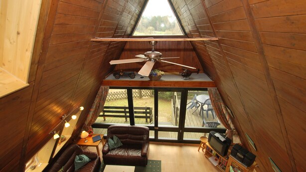 A cozy attic bedroom with a sloped ceiling, a ceiling fan, a large window, a wooden deck visible through the window, a brown leather sofa, a small table with a lamp, and a television. - Holiday park- Nurdach (Clausthal)