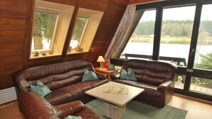 A cozy room with a brown leather sofa, a wooden coffee table, and large windows offering a view of a lake and trees. - Holiday park- Nurdach (Clausthal)