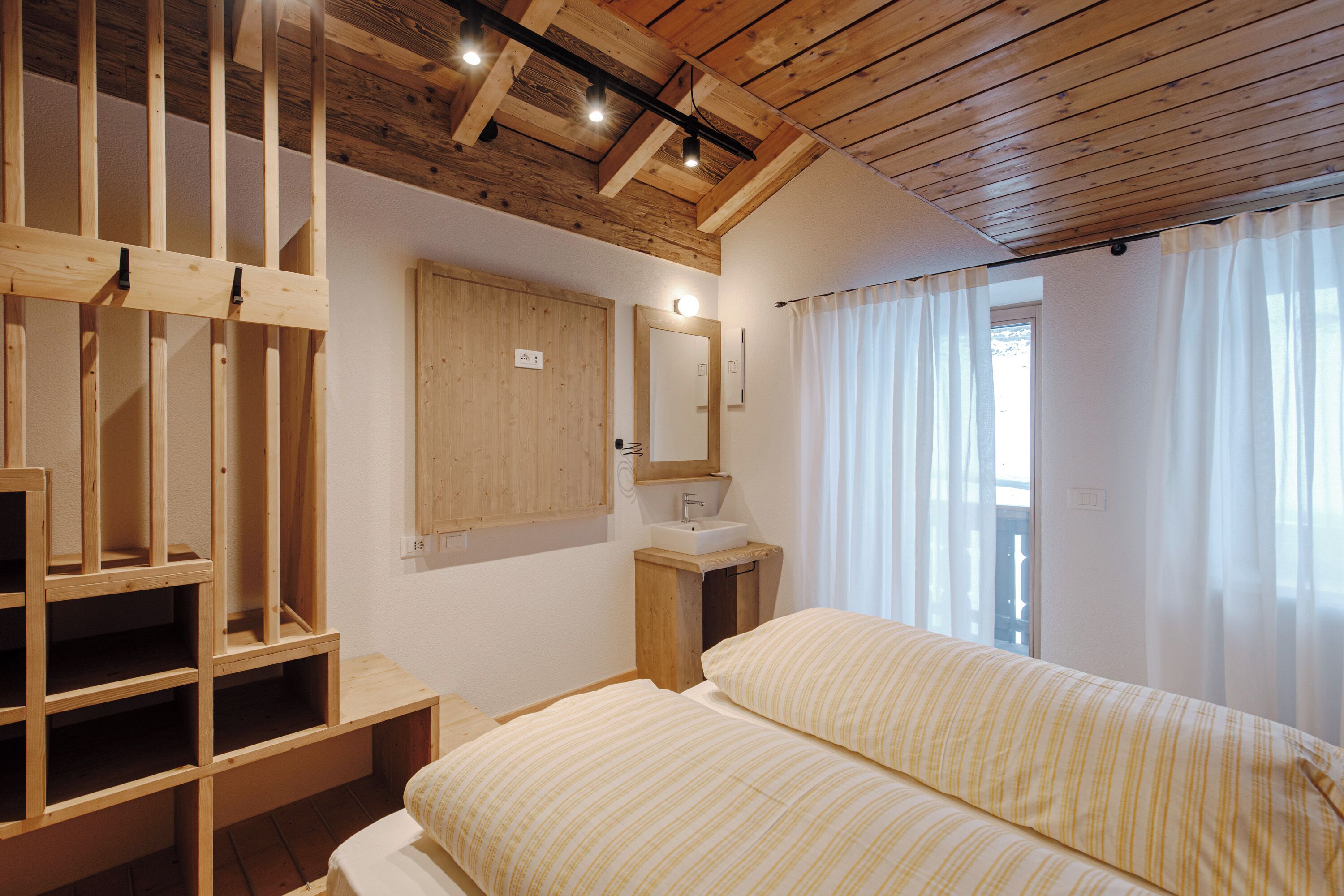 Ein Schlafzimmer mit Holzbalkendecke, einem Bett mit gestreifter Bettwäsche, einem Holzschrank, einem Nachttisch, einem Spiegel und einem Fenster mit Vorhängen.