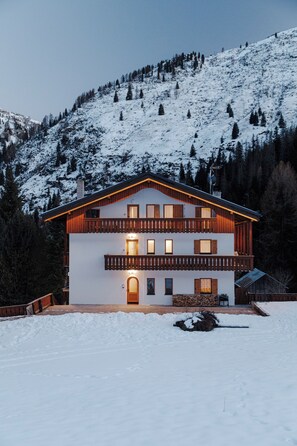 A two-story chalet with a balcony, surrounded by snow-covered mountains.