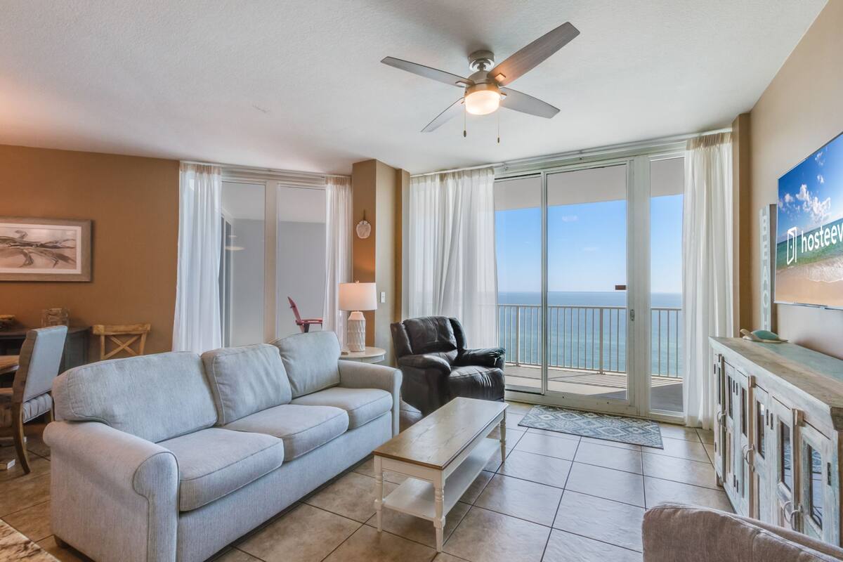 A spacious living room with a grey sofa, a black leather chair, a wooden coffee table, and a TV on the wall. A ceiling fan is visible, and the room has a large sliding glass door leading to a balcony with a view of the ocean.