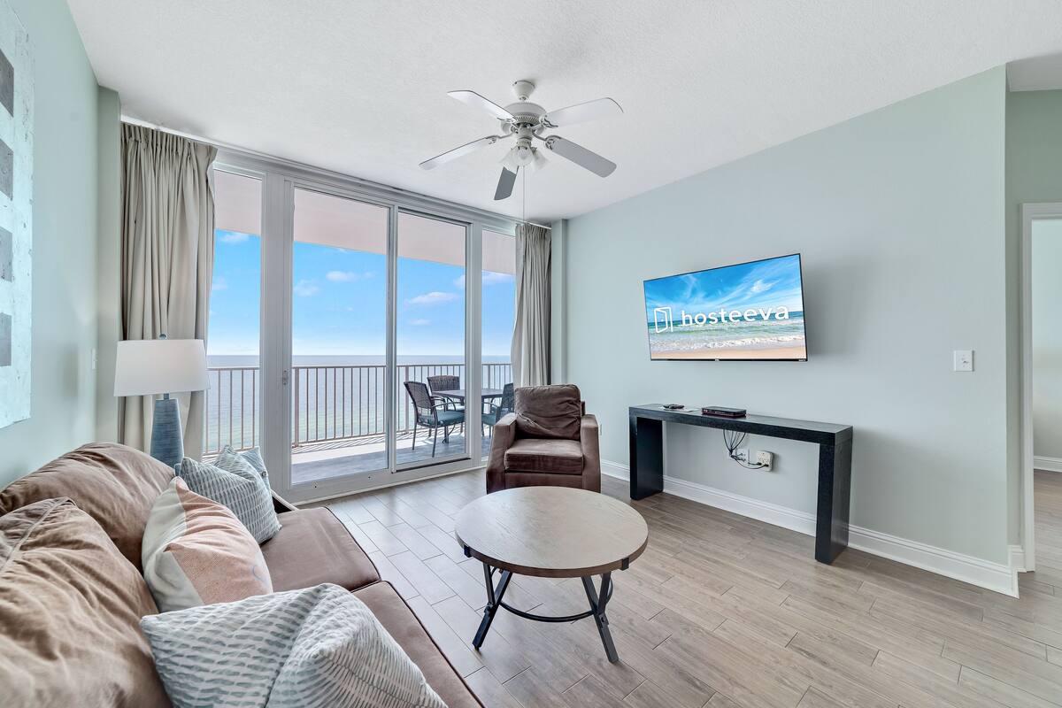 A modern living room with a large TV, a ceiling fan, a sofa, a coffee table, and a view of the ocean through a sliding glass door.