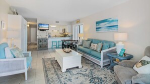A living room with a sofa, armchairs, a coffee table, and a TV. There is a kitchen in the background with a dining table and chairs. - Island Sunrise Condos by Hosteeva (Gulf Shores)
