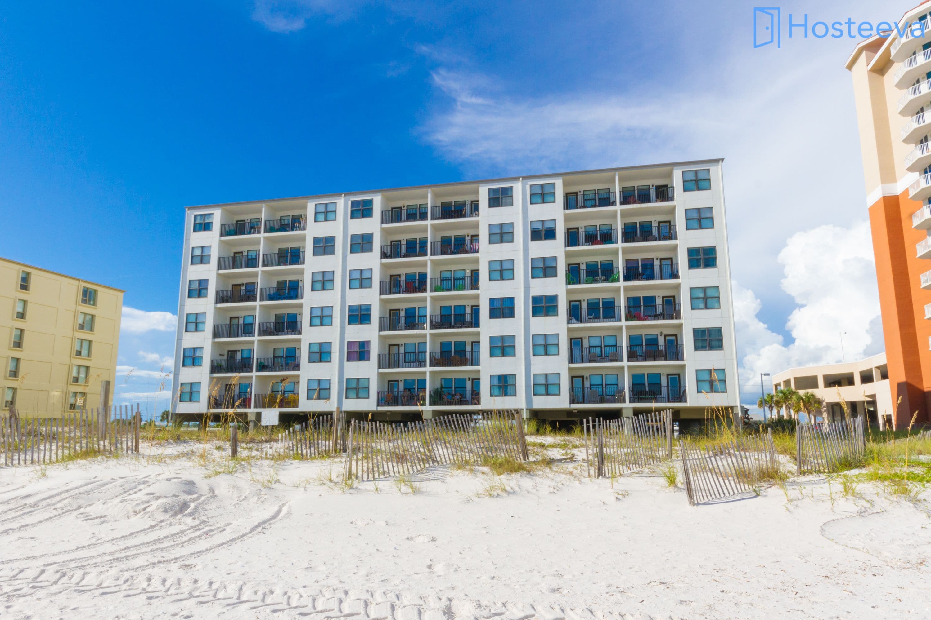 A multi-story hotel with balconies, located on a sandy beachfront with a clear sky.