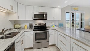 A modern kitchen with white cabinets, stainless steel appliances, and a view of the beach through the window. - Island Sunrise Condos by Hosteeva (Gulf Shores)