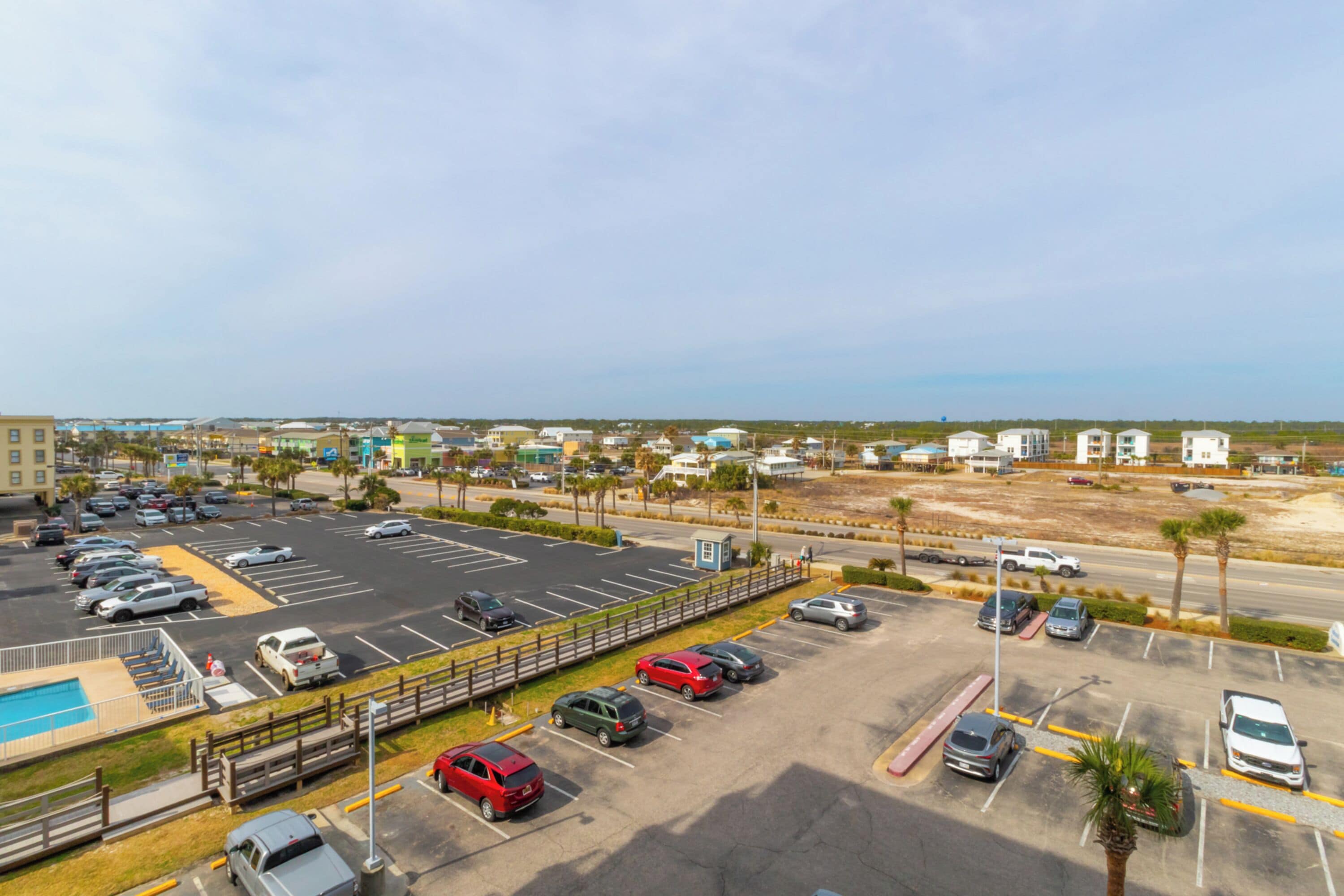 A parking lot with multiple cars, a small pool, and a view of buildings and a beach in the background.