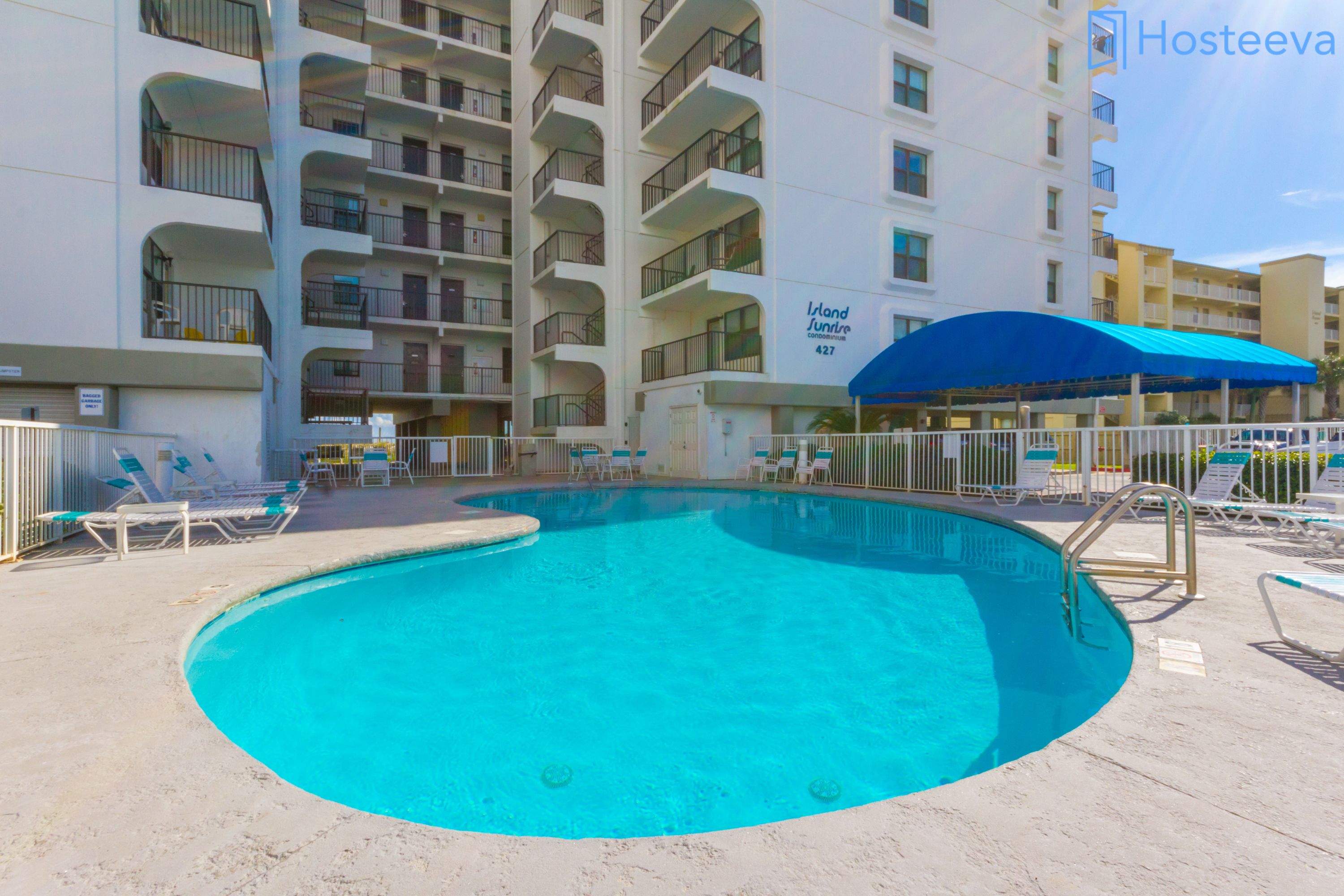 A swimming pool with lounge chairs and a blue canopy, surrounded by a multi-story building with balconies.