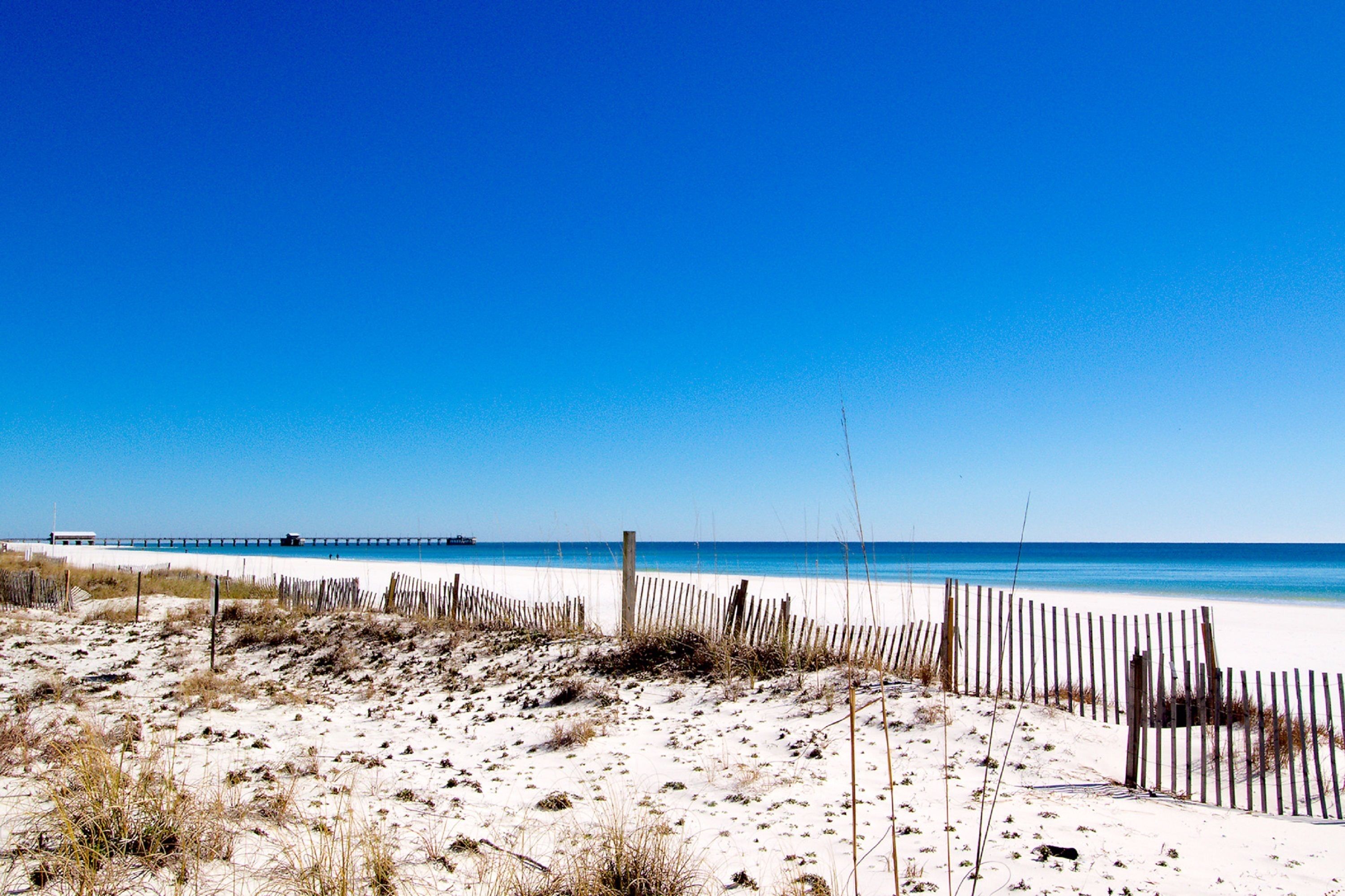 A beach with a wooden fence, sandy dunes, and a pier in the distance.
