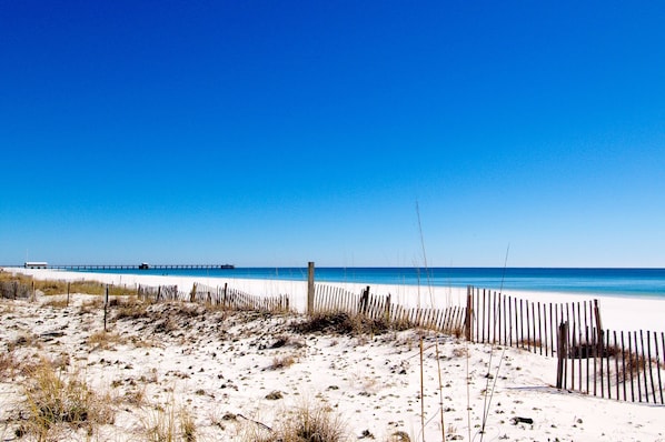A beach with a wooden fence, sandy dunes, and a pier in the distance.