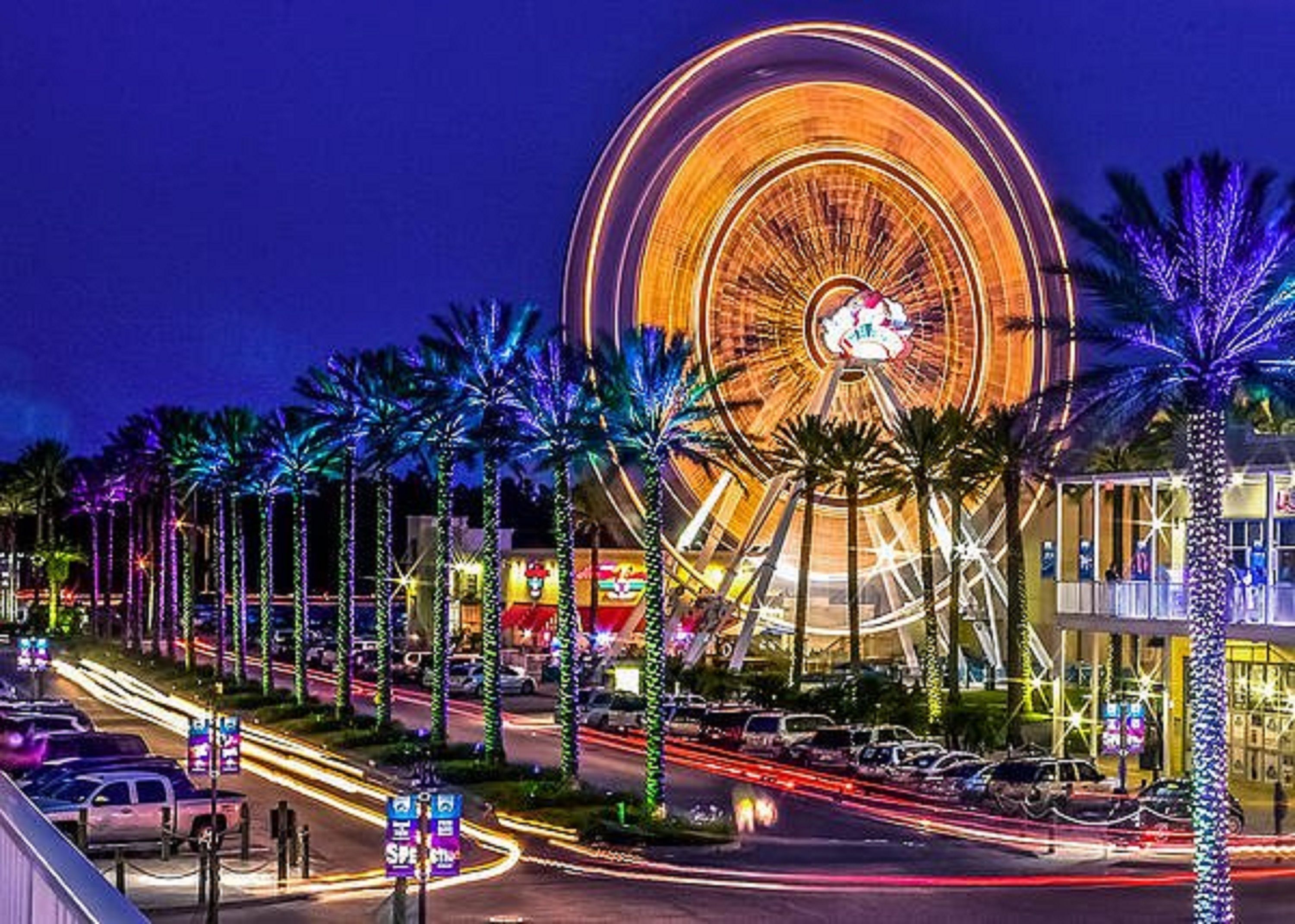 Une grande roue illuminée la nuit, avec des palmiers et des bâtiments éclairés en arrière-plan.