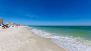Una playa con cielo azul despejado, orilla arenosa y algunos edificios al fondo.