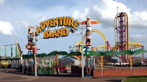 Entrada a un parque temático con un letrero que dice "Adventure Island" y varias atracciones de feria al fondo.