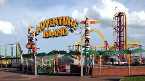 Entrada a un parque temático con un letrero que dice "Adventure Island" y varias atracciones de feria al fondo.