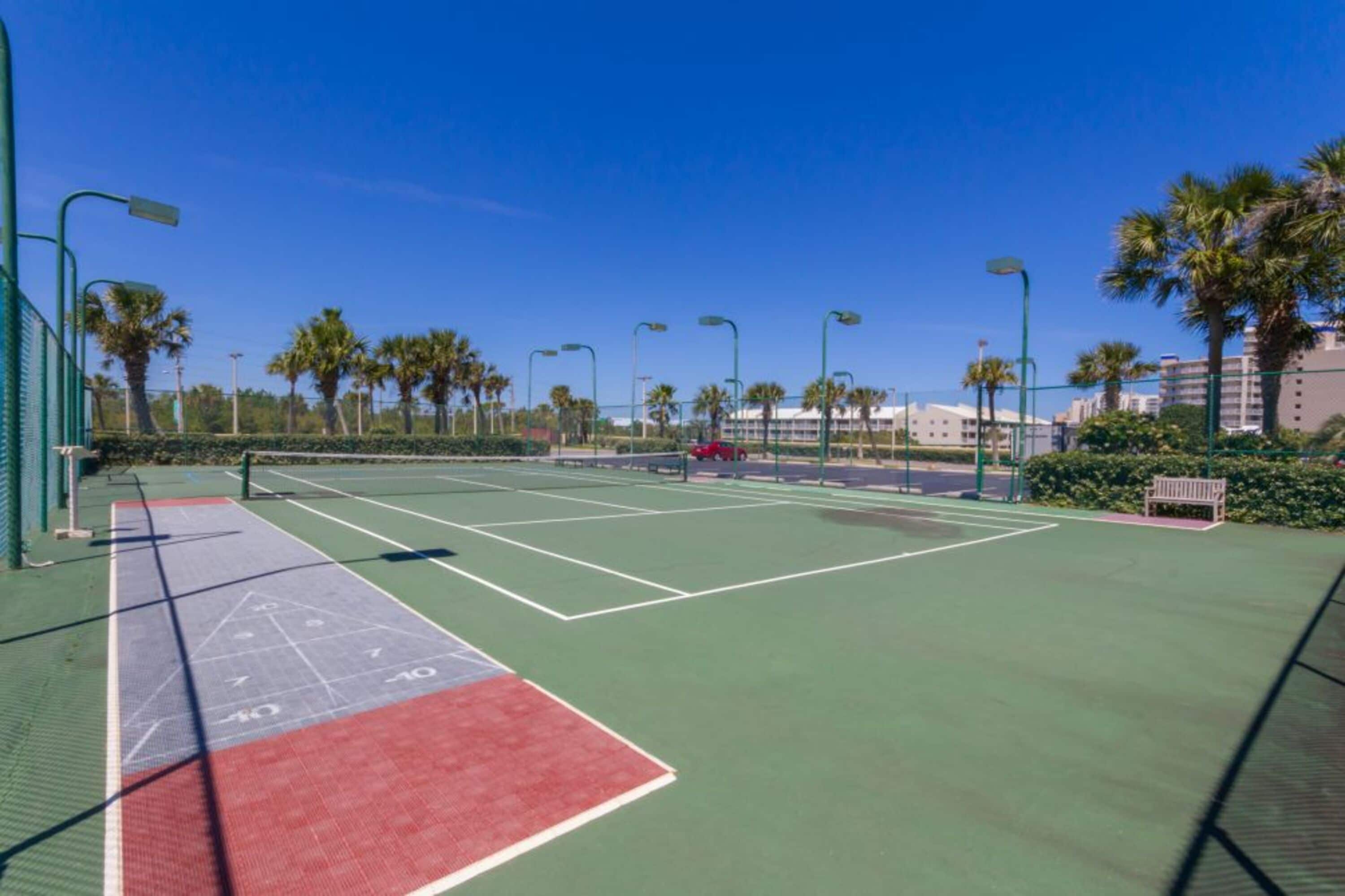 Un court de tennis en plein air avec une surface verte et rouge, entouré de palmiers et éclairé par des lampadaires.