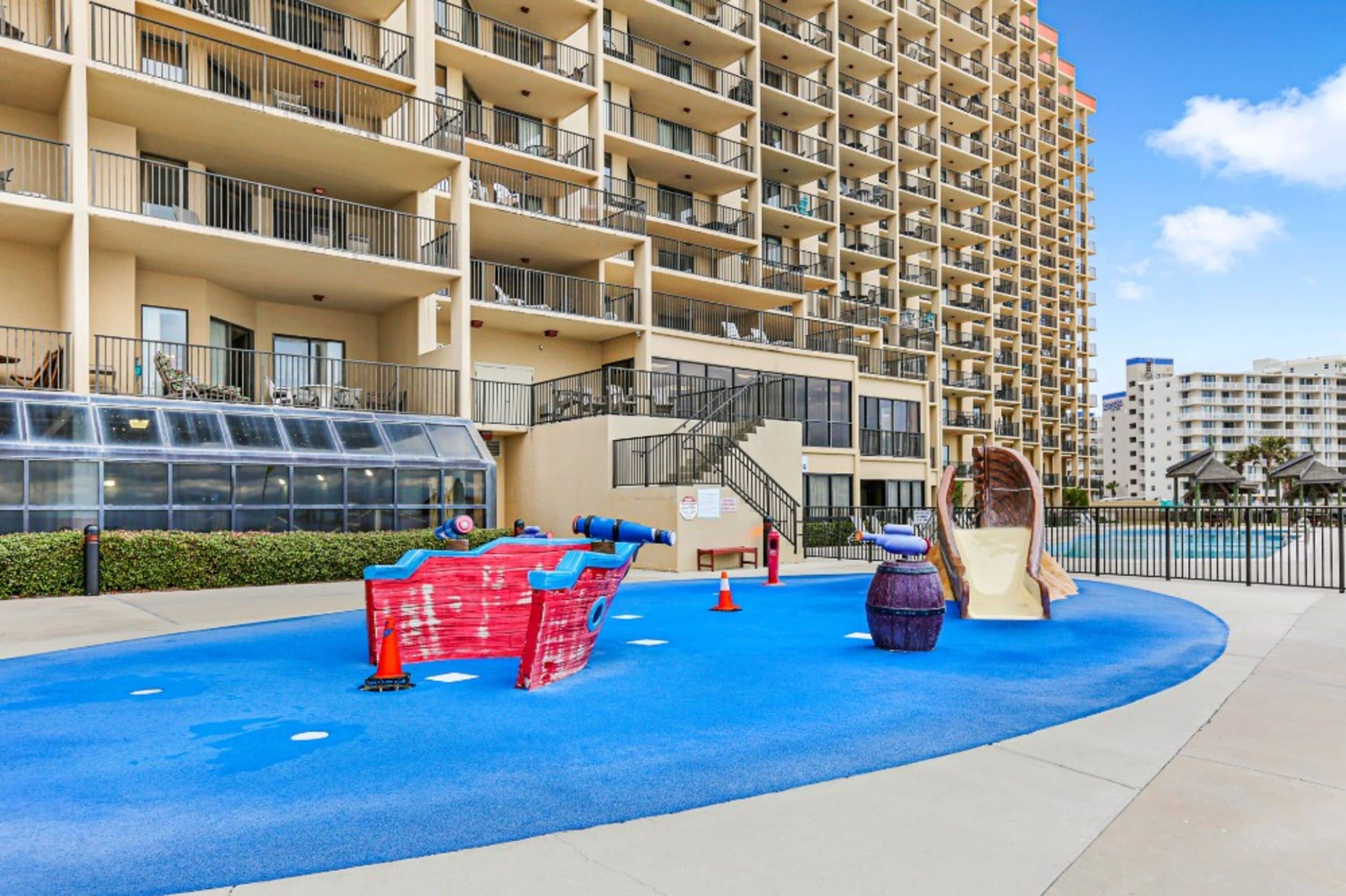 A multi-story residential building with balconies, a blue play area with slides and a small pool, and a clear sky.