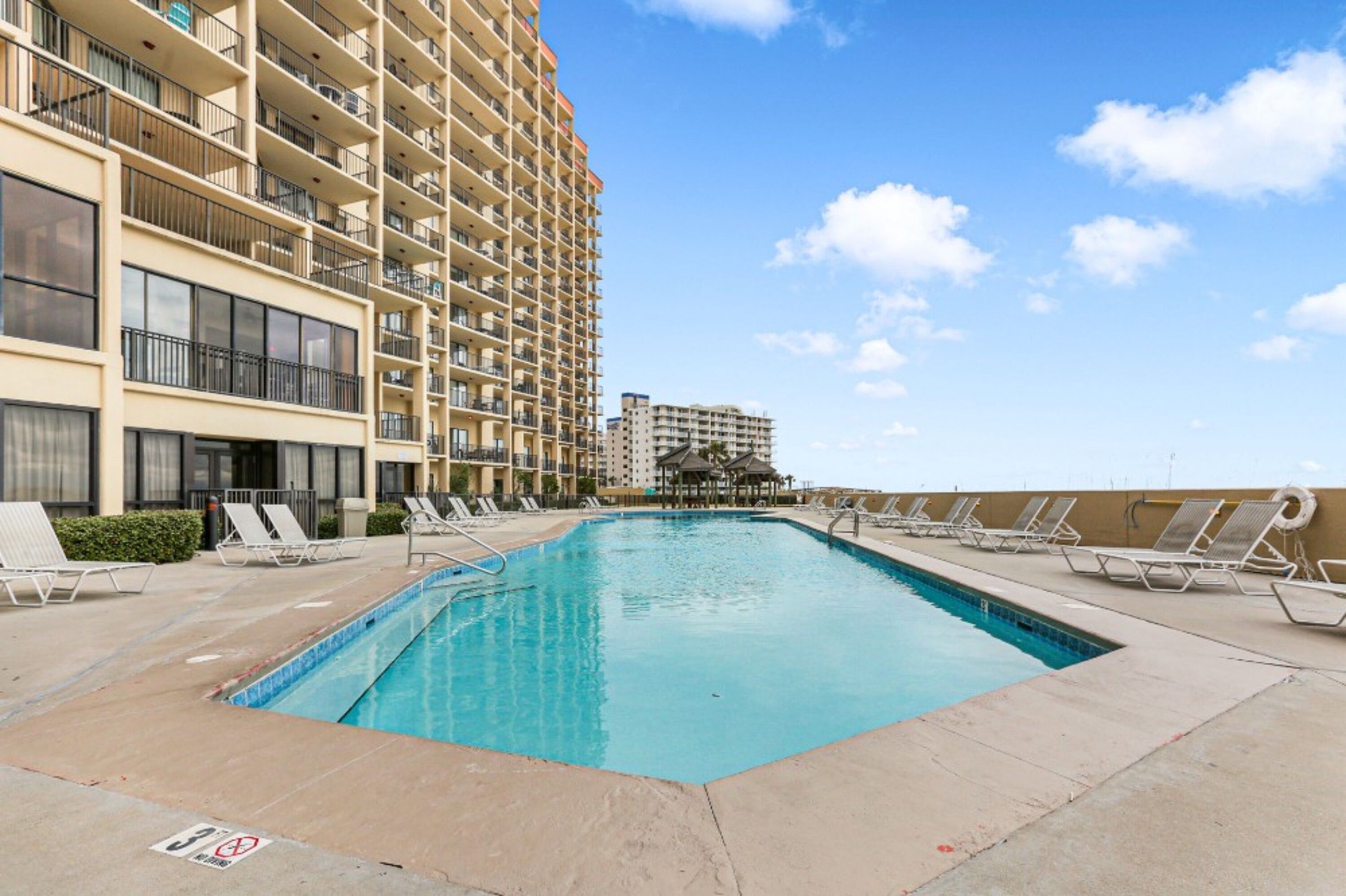Una piscina con lettini, un edificio con balconi e un cielo sereno.